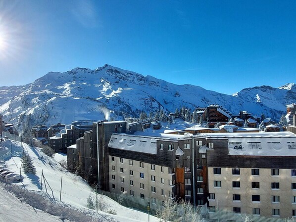 Uma cidade nas montanhas coberta de neve, com edifícios modernos, incluindo um hotel com vários andares e um teleférico visível ao fundo.