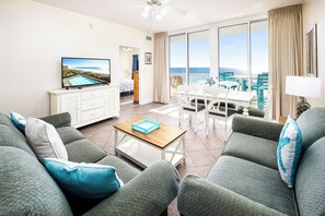 A modern living room with a large TV, a dining area, and a balcony with a view of the sea. - Waters Edge 510 • Amazing Ocean View, Balcony, Large Pool, Covered Parking, Free Beach Service (Fort Walton Beach)