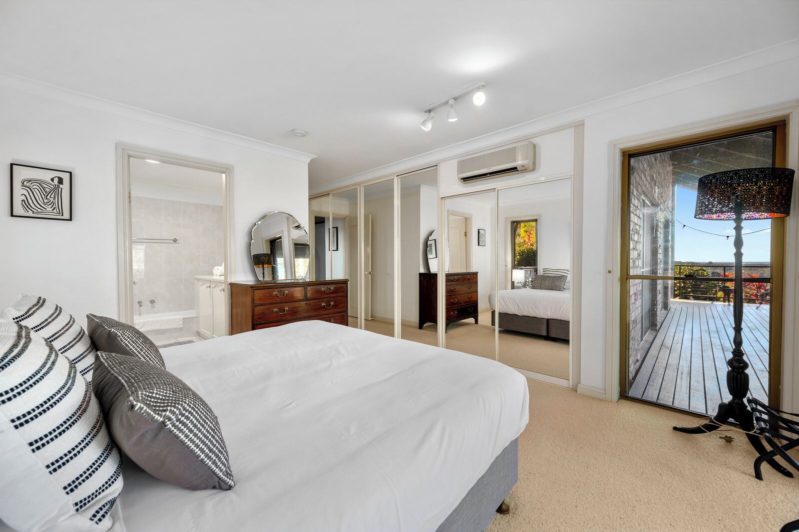 A bedroom with a large bed, a dresser, a mirror, and a view of a balcony with outdoor scenery.