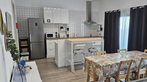 Une cuisine compacte avec des meubles blancs, un four à micro-ondes et un évier. On y trouve également un coin repas avec une table recouverte d’une nappe à motifs floraux et quatre chaises.