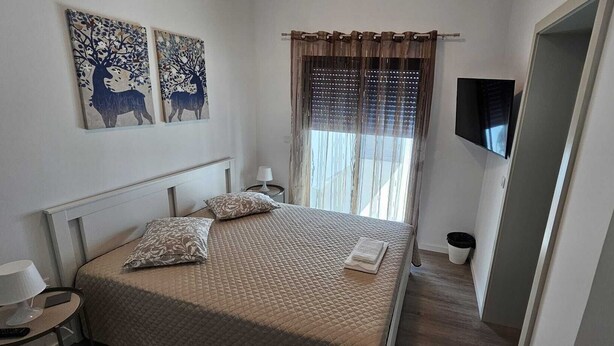 Un lit bien fait, recouvert d’une courtepointe, avec deux oreillers et une table de chevet sur laquelle se trouve une lampe. Deux tableaux sont accrochés au mur au-dessus du lit, et un téléviseur est fixé au mur.
