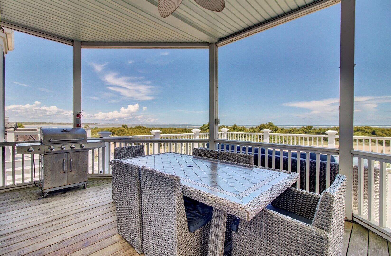 Een overdekt terras buiten met een barbecue, rieten meubels en uitzicht op de oceaan.