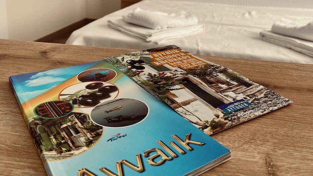 A neatly made bed with white linens, a wooden nightstand with a lamp, and a book titled "Avvalik" on a wooden table. - Noa Ayvalık Guest House (Ayvalik)