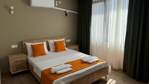 A hotel room with a wooden bed, white bedding, orange pillows, a white air conditioner, a bedside table, and a window with sheer curtains. - Noa Ayvalık Guest House (Ayvalik)