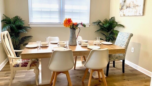 A dining area with a wooden table set for four, surrounded by white chairs. There are two potted plants on either side of the window, and a vase with flowers on the table. - Gulfport Beach Home By Casino (Gulfport)