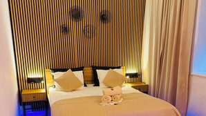 Una habitación de hotel moderna con cabecera de madera, cama con colcha de diseño romboidal, dos mesitas de noche con lámparas y una silla negra.