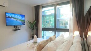 Quarto de hotel com uma cama, televisão fixada na parede, planta em vaso e vista para o exterior através de uma janela grande.
