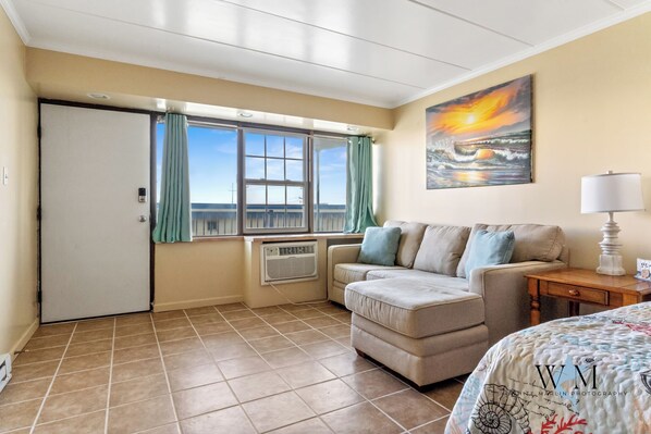 A hotel room with a sliding door, a window with curtains, a sofa, a lamp, and a painting of a sunset.
