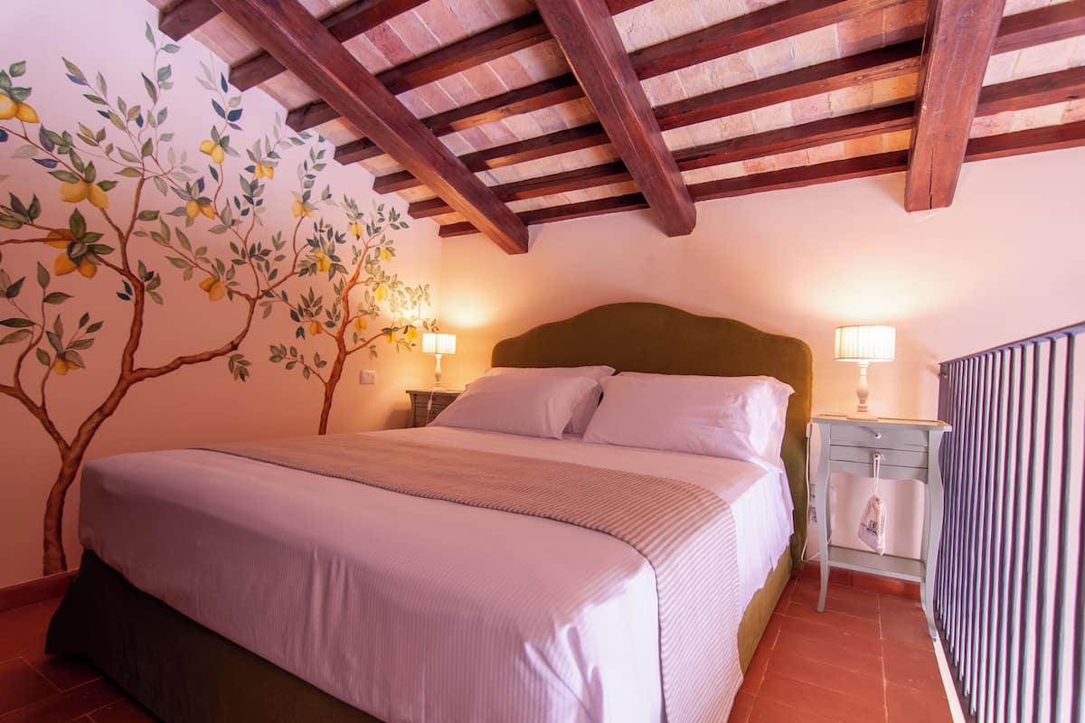 A bedroom with a large bed, wooden beams on the ceiling, a nightstand with a lamp, and a wall mural of a tree with fruit.