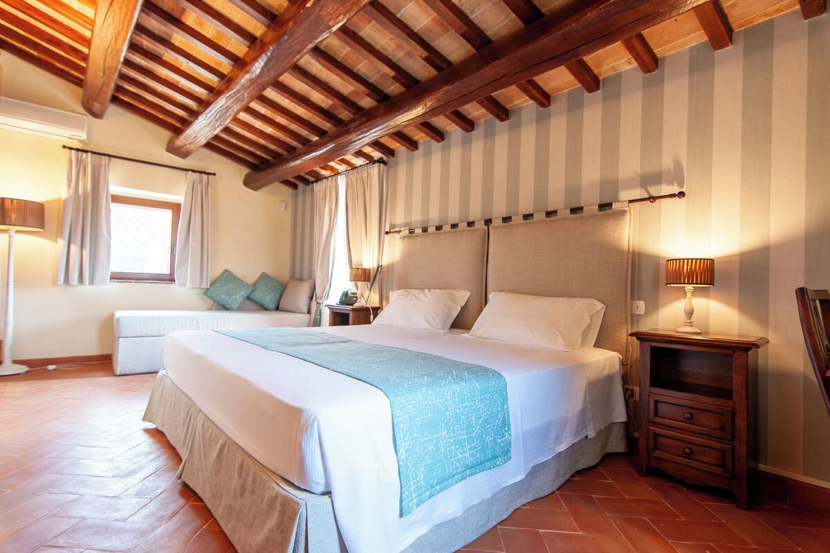 A bedroom with a large bed, wooden ceiling beams, a nightstand with a lamp, and a window with curtains.