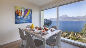 Un comedor con una mesa dispuesta para el desayuno, un cuadro de veleros en la pared, y vistas al mar y a las montañas.