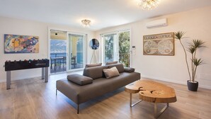 Una sala moderna con sofá, una mesa de centro y una mesa de fútbol de mesa. Hay ventanas amplias, un balcón y una planta en maceta.