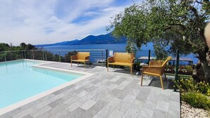 Una piscina con vistas a un lago y montañas, rodeada por un solárium de baldosas y sillas de madera.