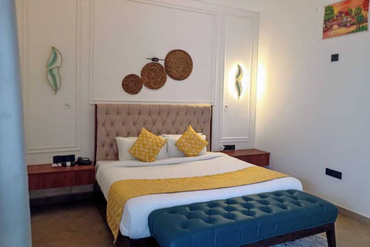 Une chambre à coucher avec une tête de lit capitonnée, un grand lit avec une literie jaune et blanche, un banc bleu et des œuvres d’art décoratives au mur.