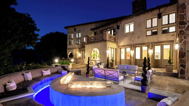 A well-lit outdoor living area with a circular seating arrangement, a fire feature, and a stone building in the background.