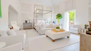 A spacious, white-themed bedroom with a canopy bed, a sitting area with a white sofa, and a large window providing natural light.