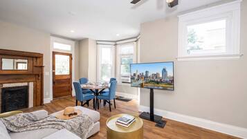 A living room with a fireplace, a dining area with blue chairs, a television mounted on the wall, and a large window.