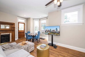 A living room with a fireplace, a dining area with blue chairs, a television mounted on the wall, and a large window. - Near Cincy Skyline Views Historic 4BR (Covington)