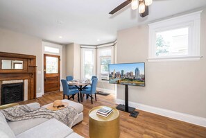 A living room with a fireplace, a dining area with blue chairs, a television mounted on the wall, and a sofa with a hat and sunglasses on it. - Near Cincy Skyline Views Historic 4BR (Covington)