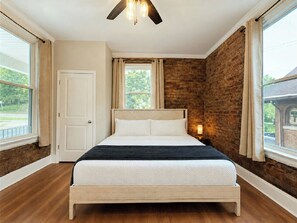 A bedroom with a brick wall, a wooden bed, a ceiling fan, and a window with curtains. - Near Cincy Skyline Views Historic 4BR (Covington)