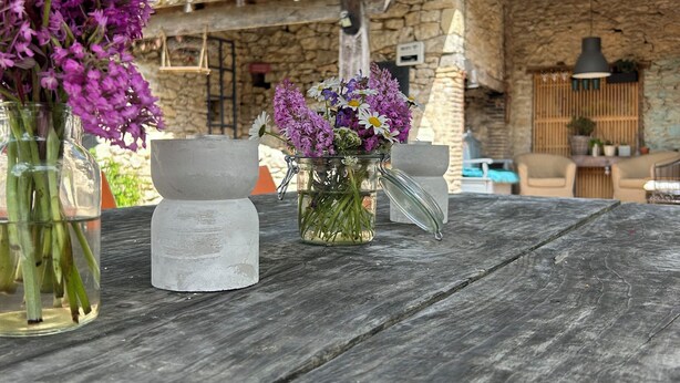 Un décor extérieur rustique avec un bâtiment en pierre, une table en bois et deux vases en verre ornés de fleurs.