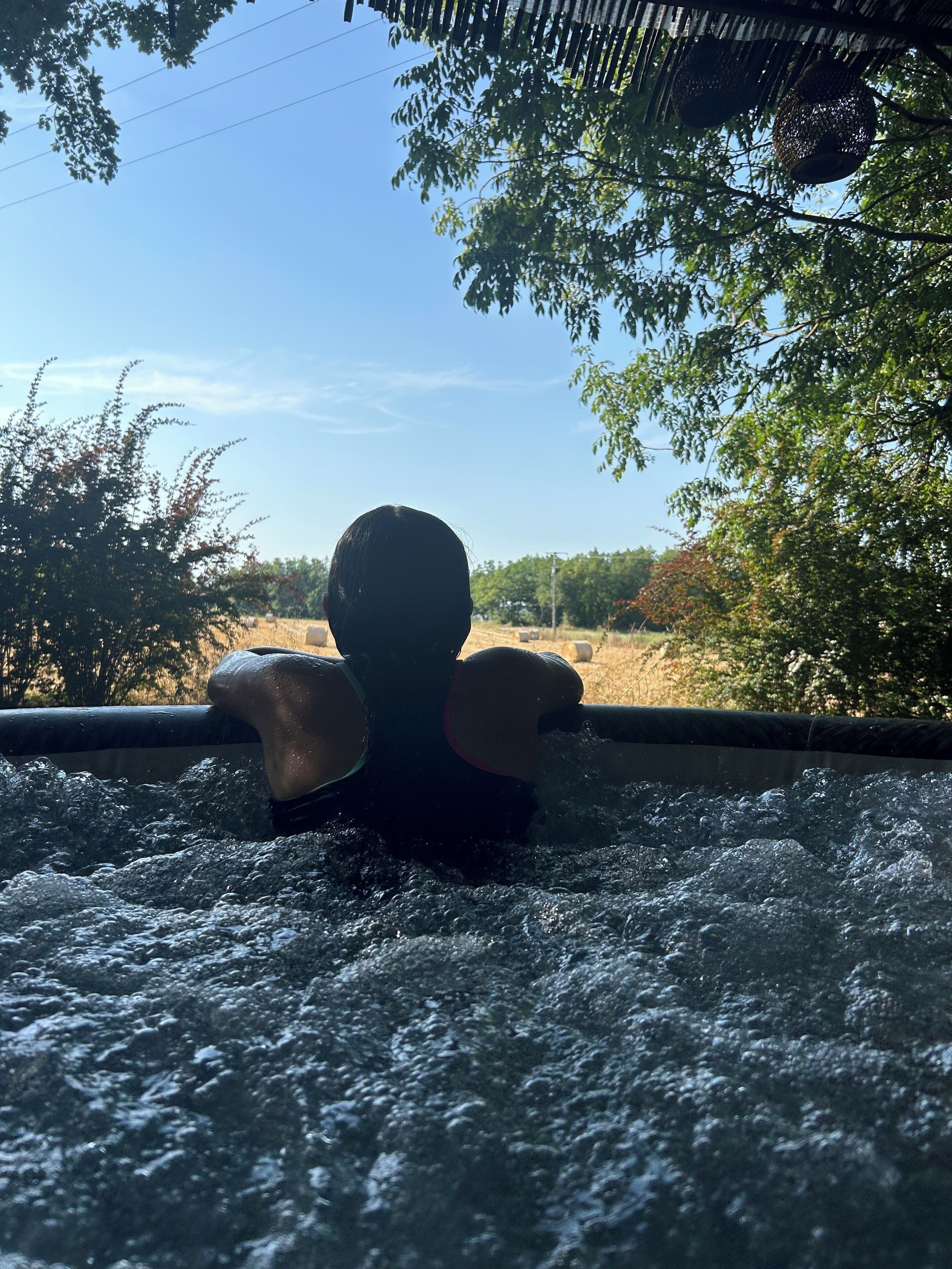 A person in a hot tub with water splashing around them, surrounded by trees and a clear sky.