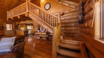 Interior de una cabaña de troncos con una escalera de madera, un comedor y una sala de estar con un sofá de cuero.