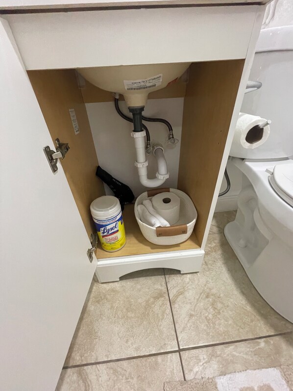 Un baño con inodoro, lavamanos de cuenca blanca, un aerosol desinfectante Lysol y un dispensador de papel higiénico blanco.