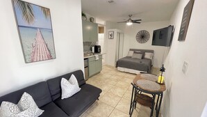 Un espace de vie compact comprenant un canapé, une petite table ronde, un lit, un coin cuisine et un ventilateur de plafond.