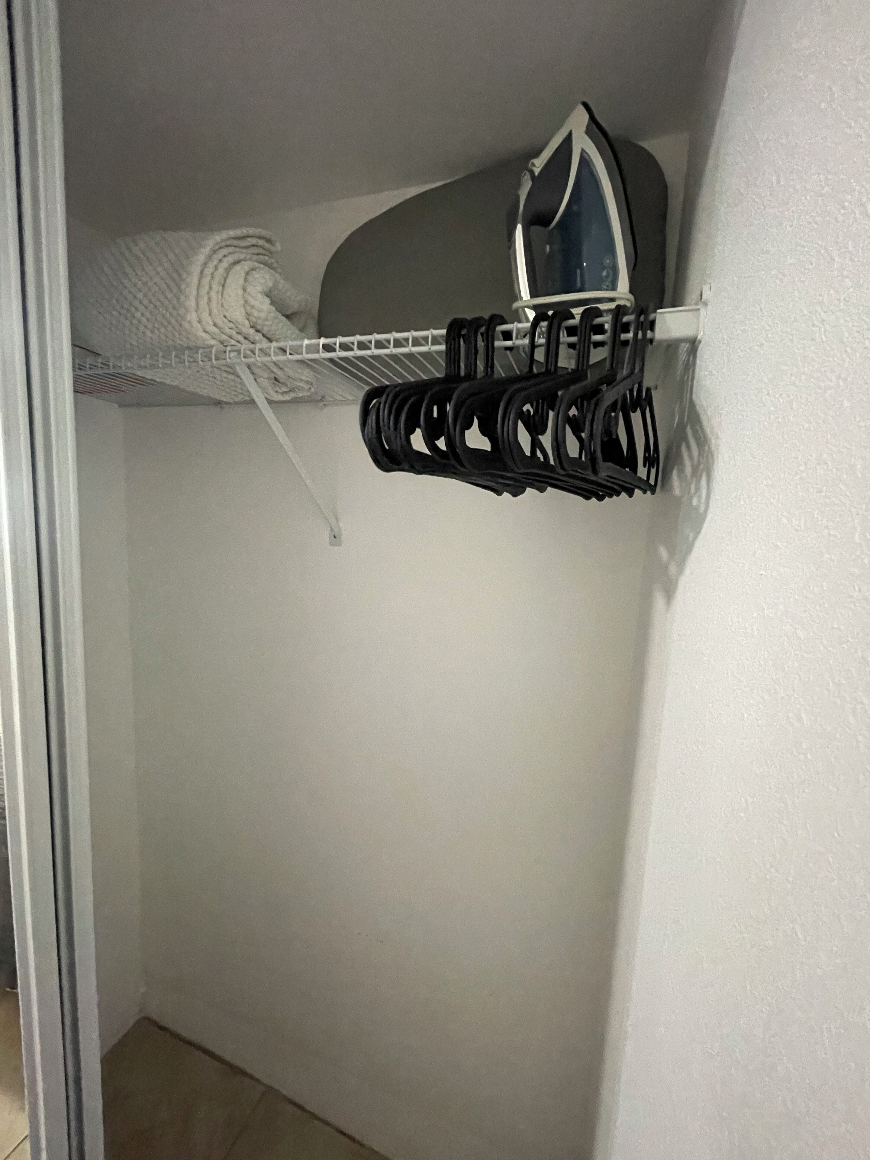 A storage closet with a white shelf holding a folded towel and a black hanging rack with clothes.