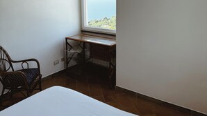 Ein kleiner, heller Raum mit einem Bett, einem Korbstuhl und einem Schreibtisch mit Meerblick.