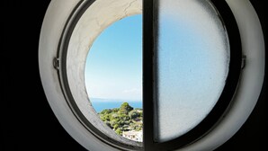 Ein rundes Fenster mit freiem Blick auf eine Küstenlandschaft, darunter Grünpflanzen und ein ferner Horizont.