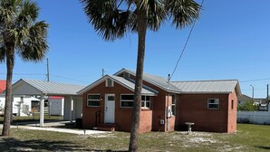 A small brick building with a white door and windows, surrounded by a fenced yard and a palm tree.