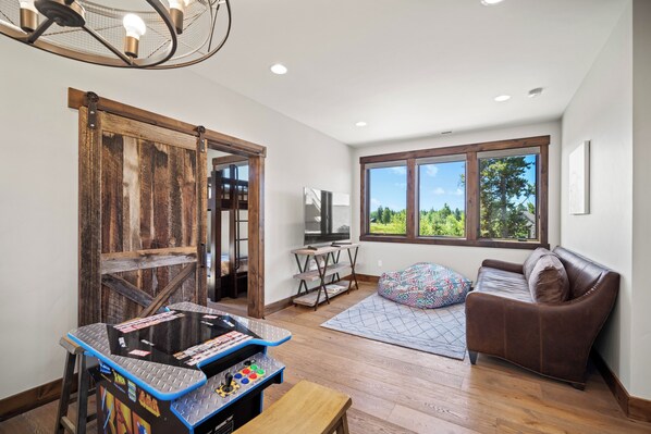 A modern living room with a wooden floor, a brown leather sofa, a gaming table, and a large window offering a view of trees. - Golf Course Escape with Access to Exclusive Golf (Bend)