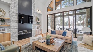 A spacious living room with a stone fireplace, large windows, and a high ceiling. - Luxury Family Retreat with Access to Exclusive Golf (Sunriver)