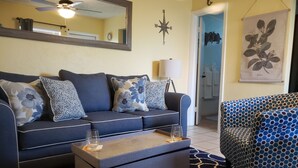 Un salon comprenant un canapé, une table basse, un fauteuil à motifs, une horloge murale, un ventilateur de plafond, un miroir et une boussole fixée au mur.