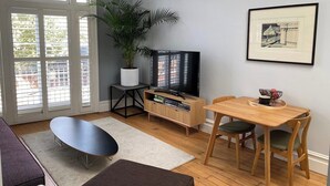Ein modernes Wohnzimmer mit Fernseher, Esstisch aus Holz und einem großen Fenster mit Jalousien.
