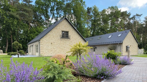 Ein Steinhaus mit einem geneigten Dach, umgeben von einem gepflegten Garten mit violetten Blumen und grünen Sträuchern.