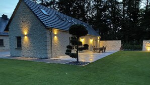Ein Steinhaus mit geneigtem Dach, ein kleiner Baum im Vordergrund und eine Terrasse mit Gartenmöbeln.