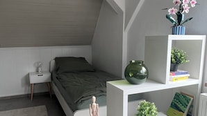 Een slaapkamer met een schuine plafond, een bed met een groene dekbedovertrek, een nachtkastje met een lamp, een klein houten figuurtje, een witte plank met boeken en planten, en een raam met luiken.