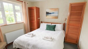 A neatly made bed with white linens, a green pillow, and two folded towels. There is a painting on the wall, a window with curtains, and a radiator under the bed. - BEN GOWER, pet friendly, with open fire in Oughterard, County Galway (GAILLIMH)