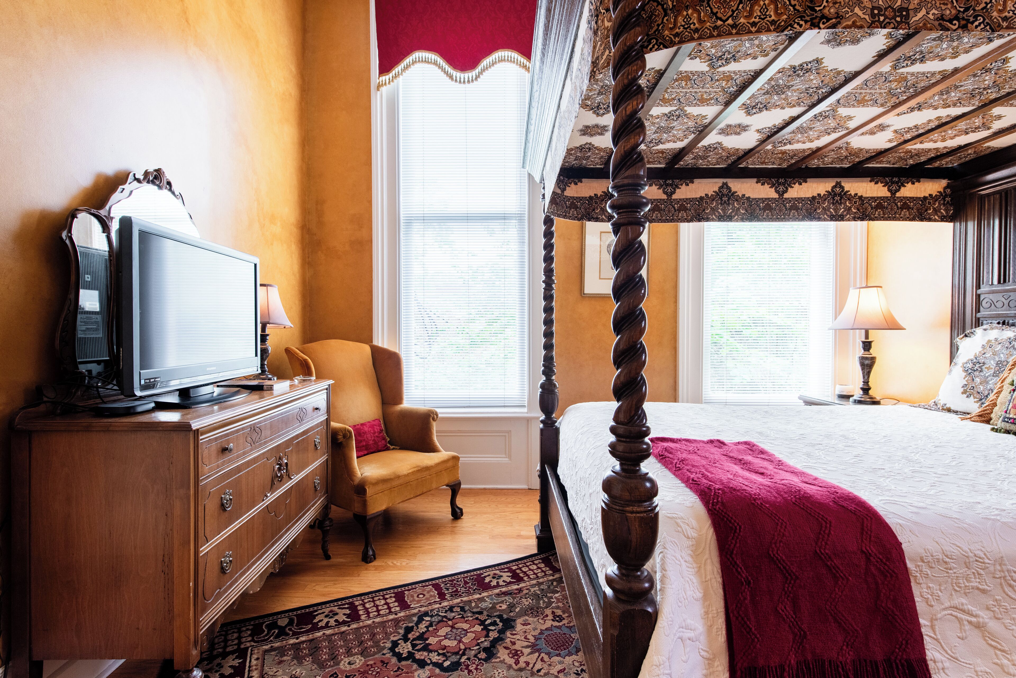 Una cama con dosel y tablero de madera, un televisor sobre una cómoda de madera, una silla y una alfombra estampada.