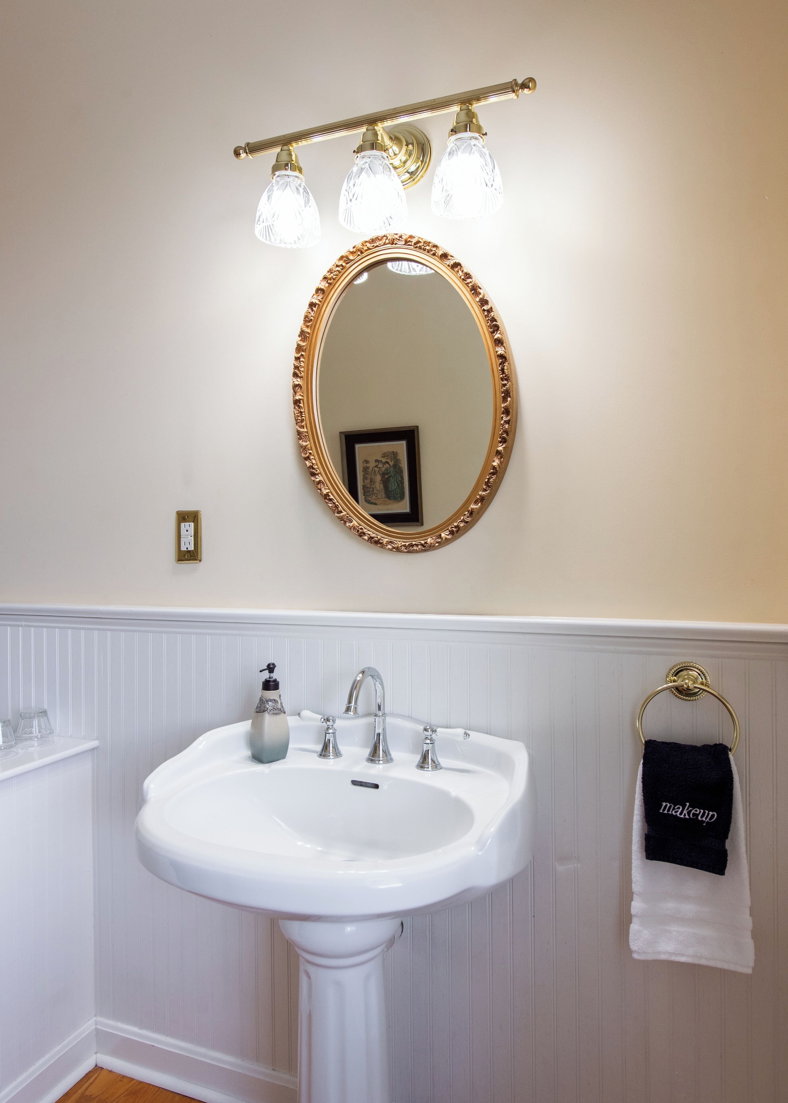 A bathroom with a white pedestal sink, a gold-framed mirror, and a vanity with a gold towel ring.