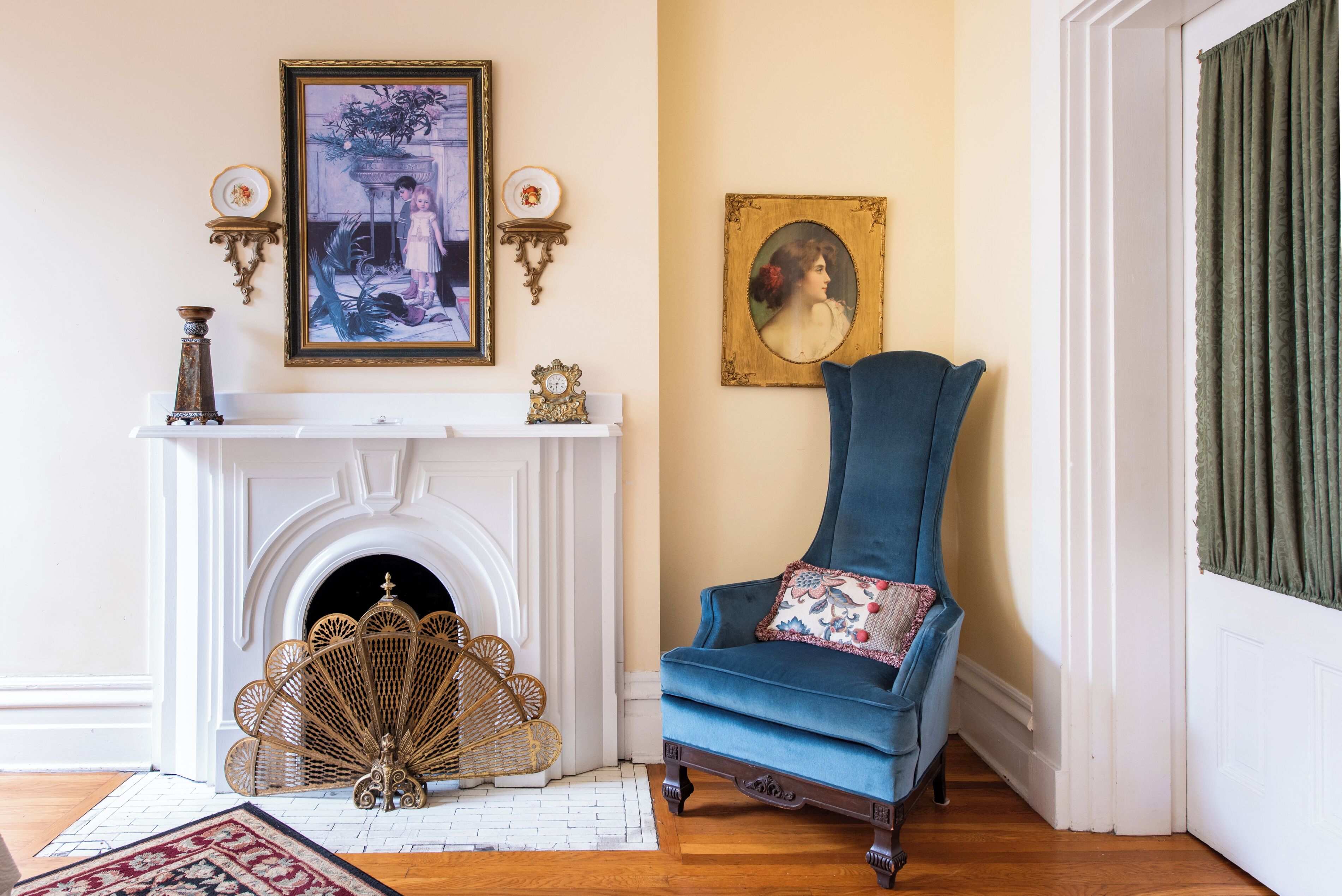 Quarto com lareira branca, poltrona azul com almofada floral e um retrato emoldurado na parede.