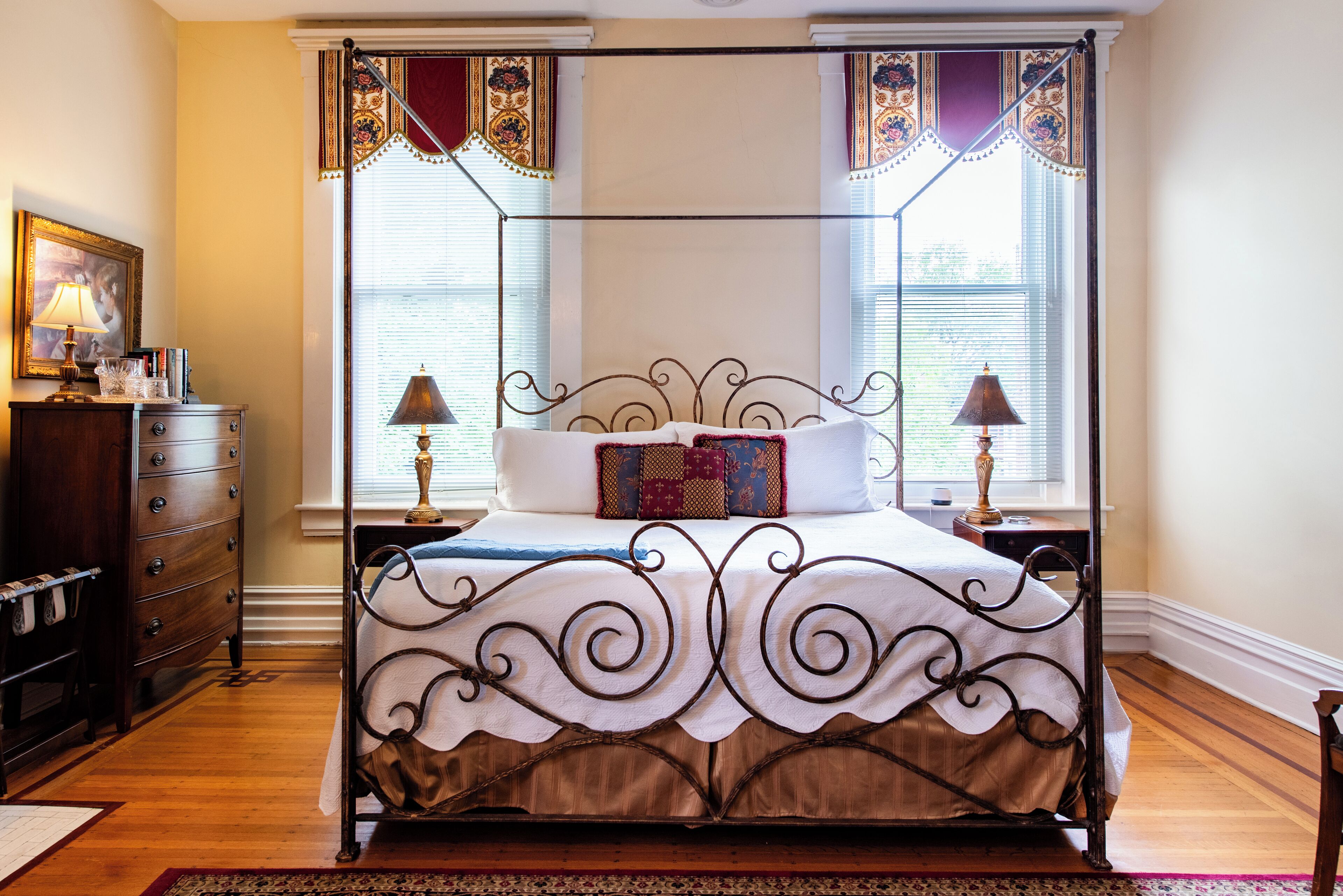 A four-poster bed with a decorative headboard, two bedside tables with lamps, a dresser with a mirror, and a window with curtains.