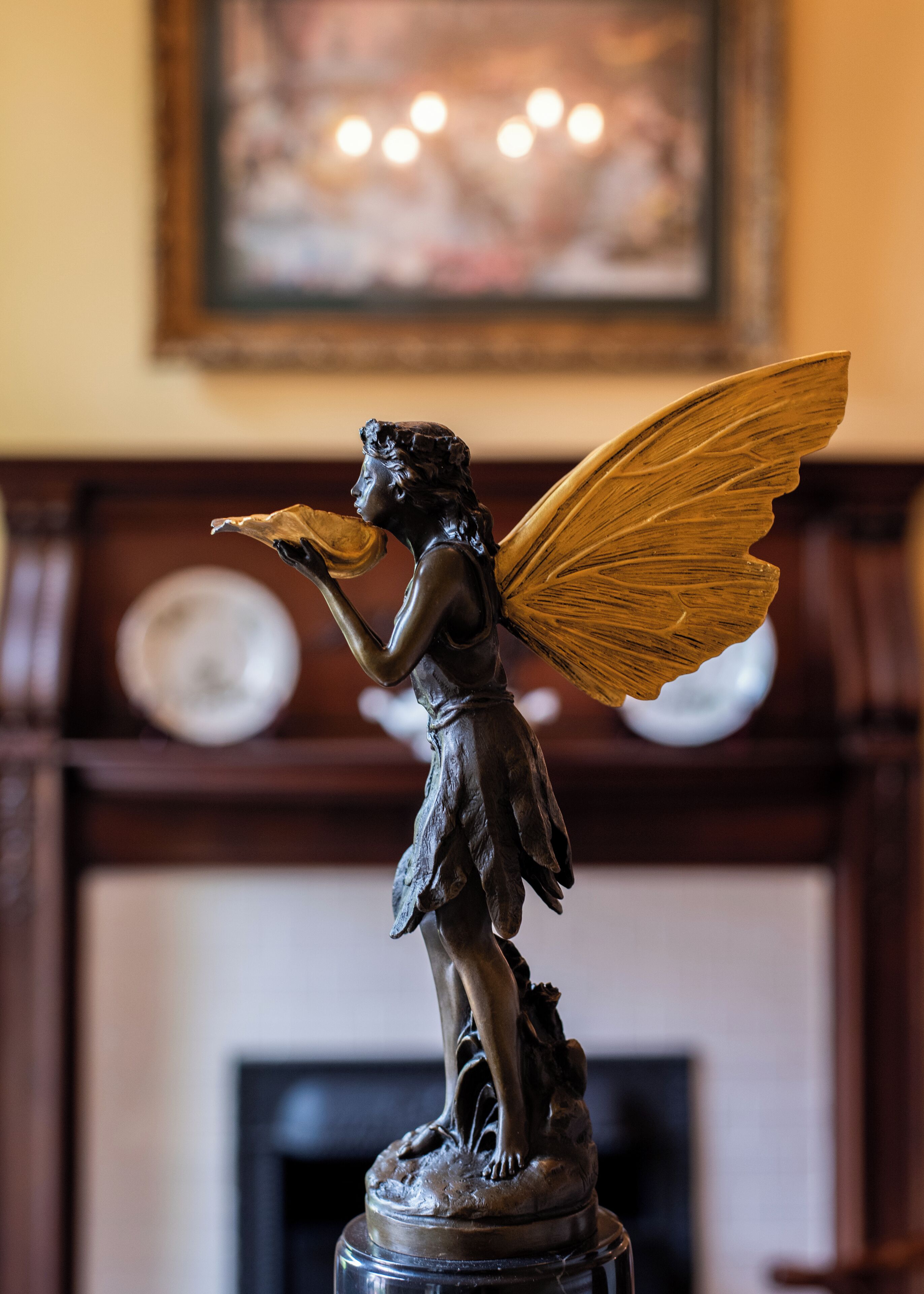 A statue of a winged figure with a book, standing on a pedestal, in a room with a fireplace and a framed picture on the wall.
