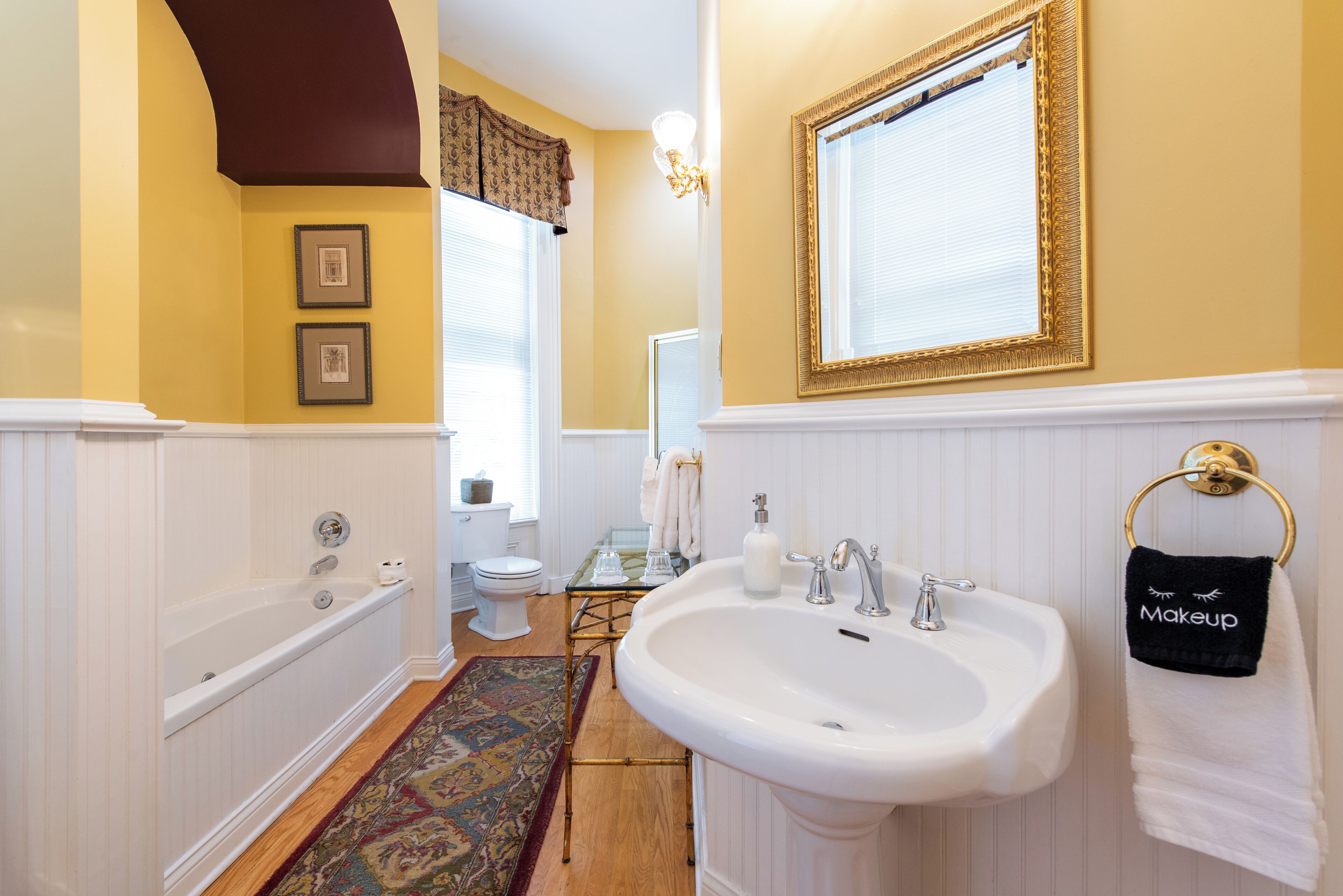 Une salle de bain avec une baignoire, un lavabo et des toilettes. Des tableaux encadrés ornent le mur, un tapis recouvre le sol et un porte-serviettes est fixé au mur, sur lequel est posée une serviette.