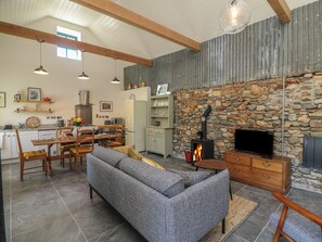 Uma sala de estar ampla com lareira de pedra, um sofá e uma televisão. Uma cozinha pode ser vista ao fundo, com uma geladeira e um micro-ondas.