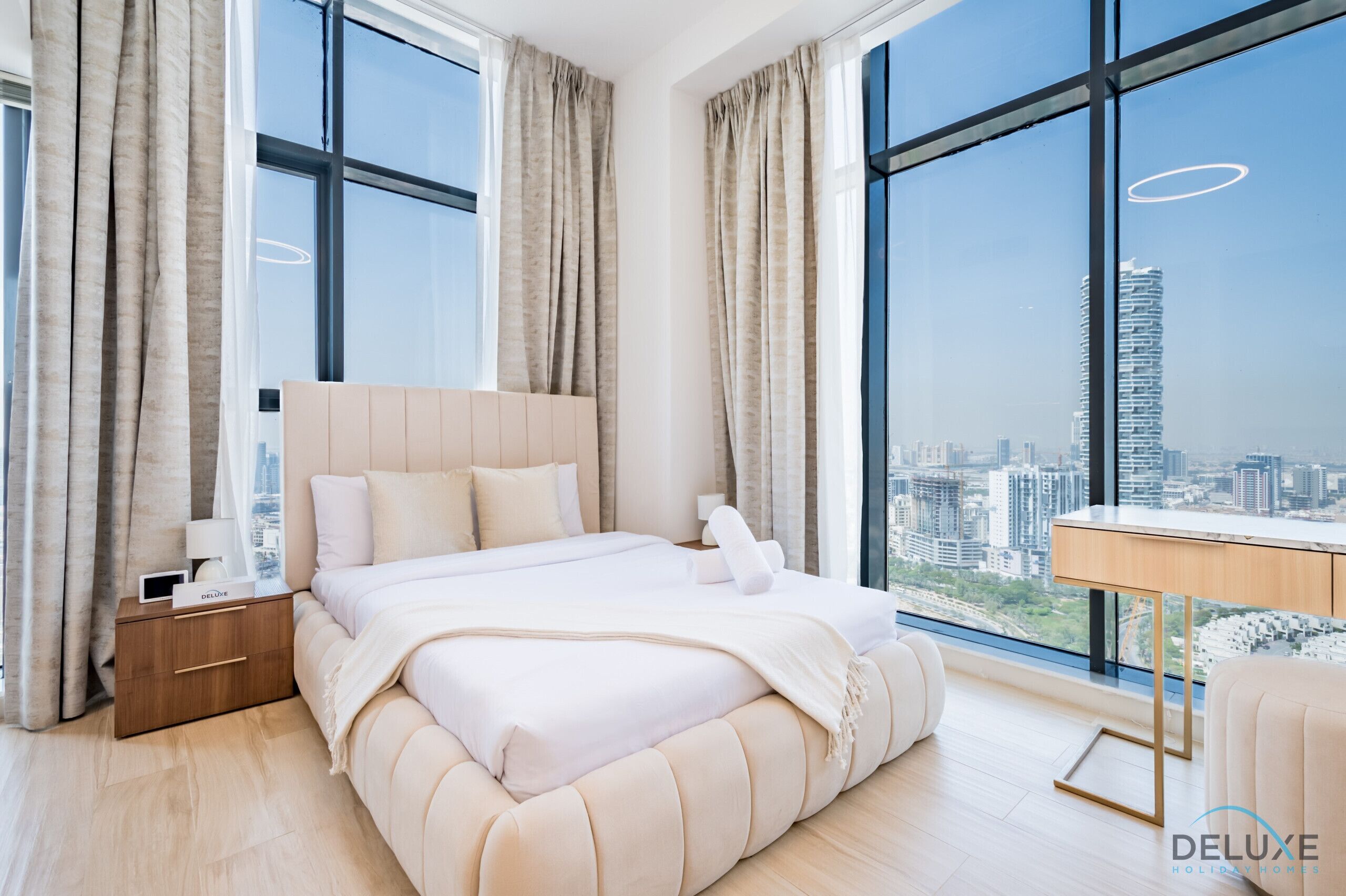 Une chambre moderne avec un grand lit, une table de chevet, un bureau et une vue sur le paysage urbain grâce à de grandes fenêtres.
