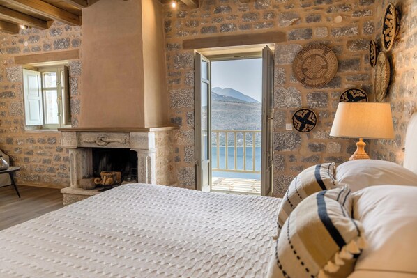 A bedroom with a stone fireplace, a bed with striped pillows, and a view of the sea through an open door.